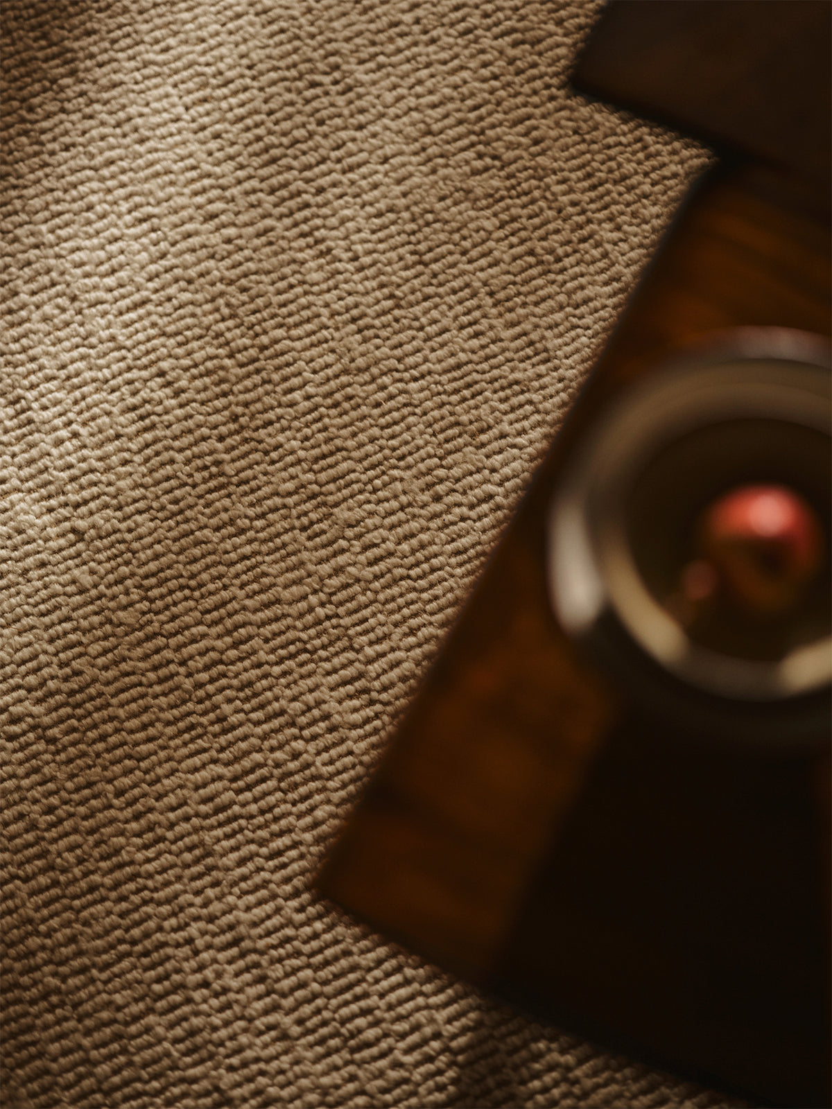 A close-up of a textured beige carpet with a dark wooden surface and a blurred round object in the foreground | Armadillo Abbey Rug