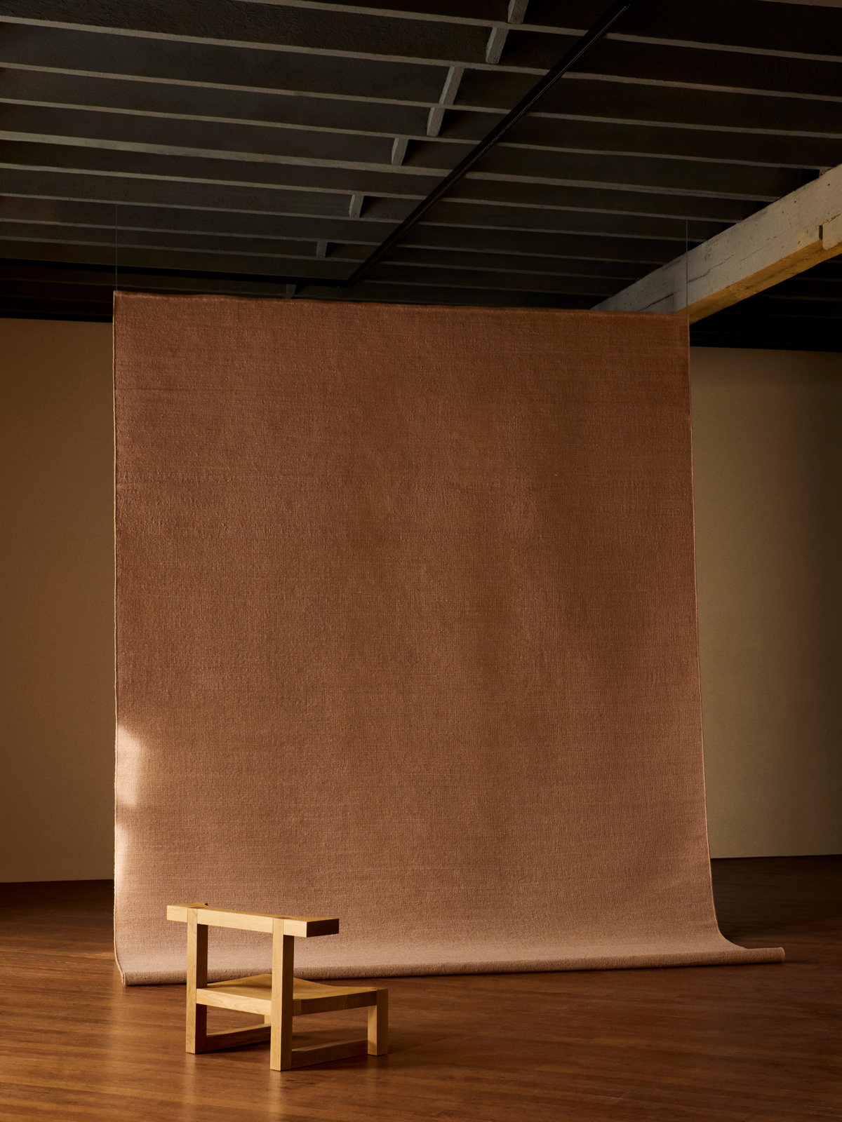 A large, textured brown rug backdrop hanging in a studio space with a wooden floor and ceiling, with a small wooden stool in the foreground | Armadillo Aerie Rug